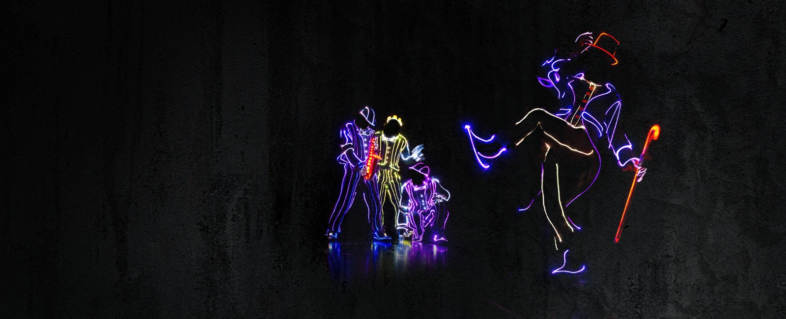 A dark background and stage are lit up by people wearing wireless LED suits of purple, red and yellow. Three people crouch on the left appearing to play instruments like the saxophone, while one person on the right is standing and kicking while appearing to wear a tophat and hold a cane.