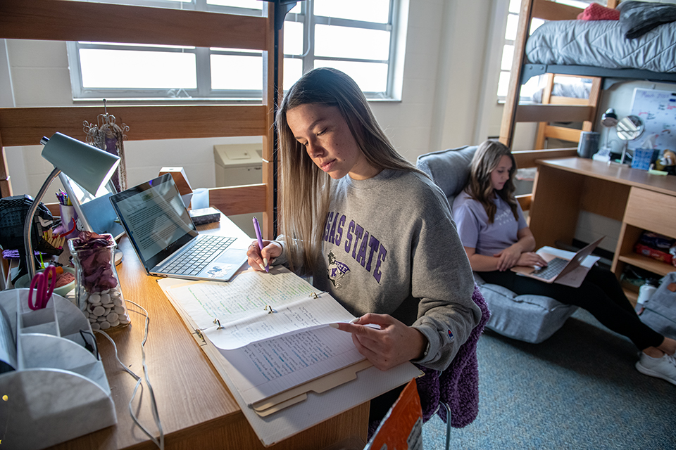 A young college student and her roommate study inside their dorm. One is at her desk, while the other is laying on the floor.