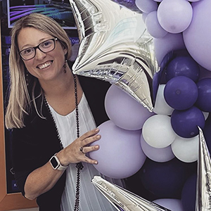 Jana Thomas, smiling big and surrounded by purple, white and silver ballons