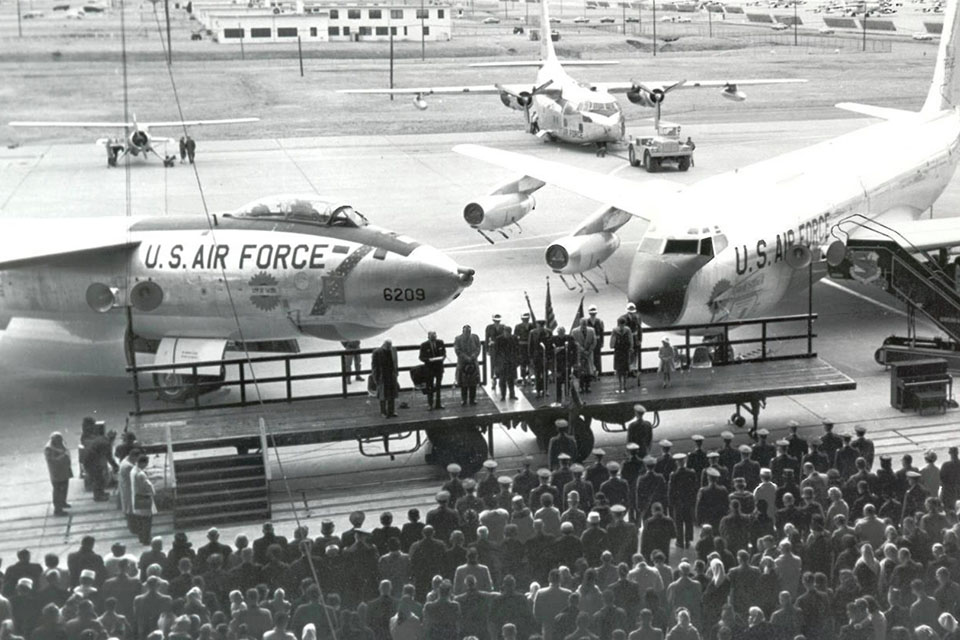 An above head view shows several crowds gathered in a hangar, with U.S. Air Force planes parked just outside the hangar doors.