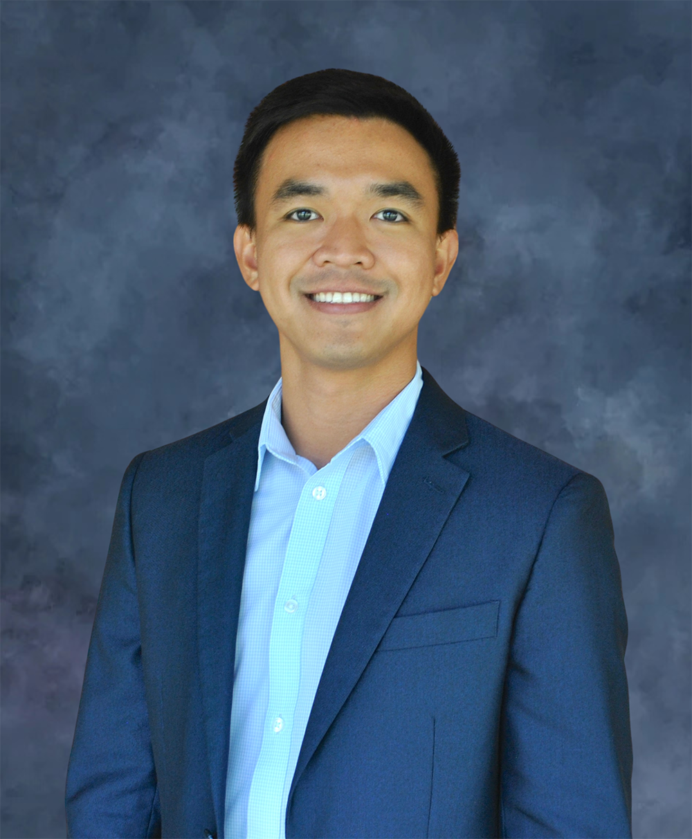 A doctoral student in a blue blazer and light blue shirt smiles against a dark gray-blue background.