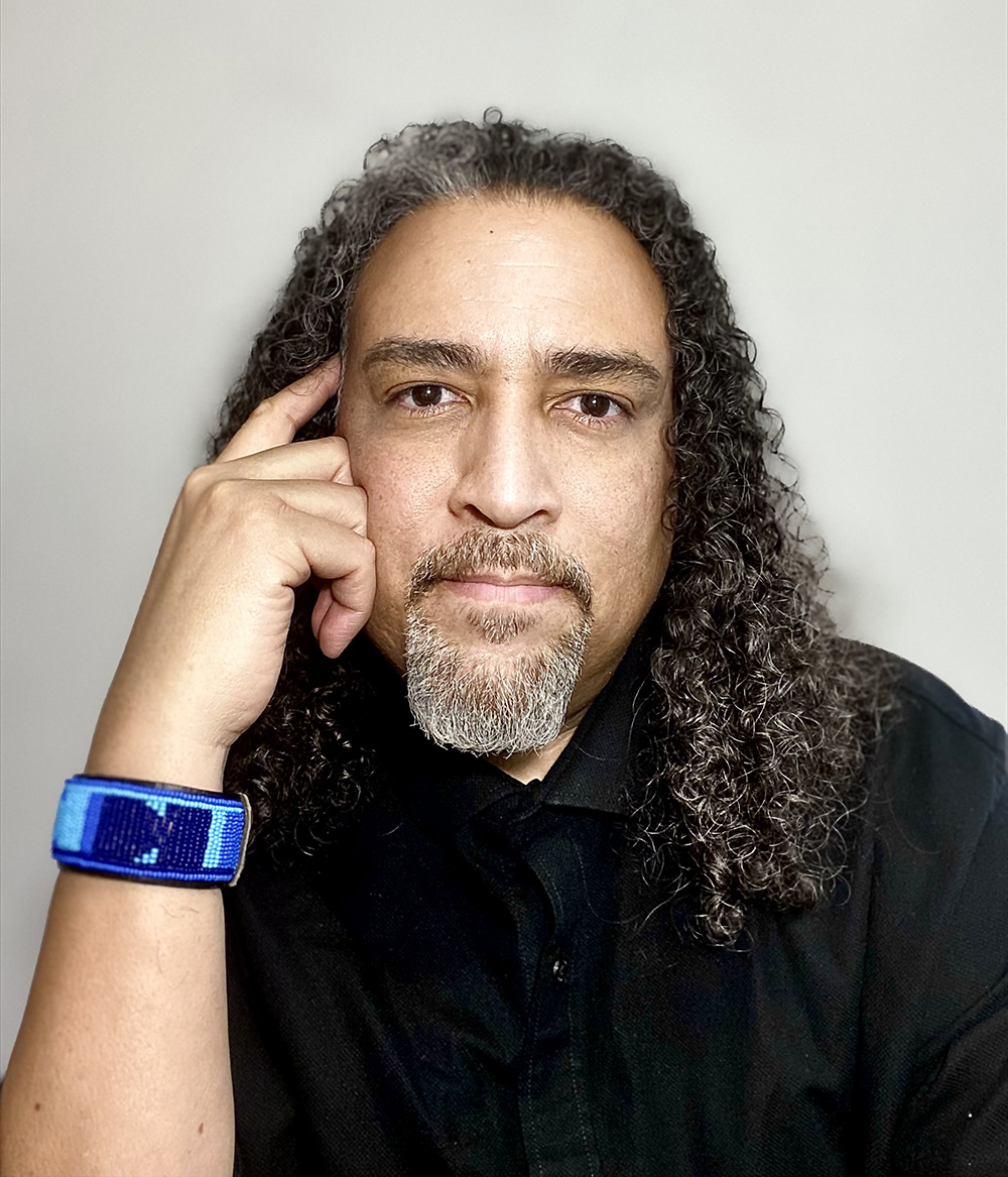A man with long, curly dark hair wearing a black button-up shirt and a blue beaded bracelet looks directly into the camera with his finger against his right temple.