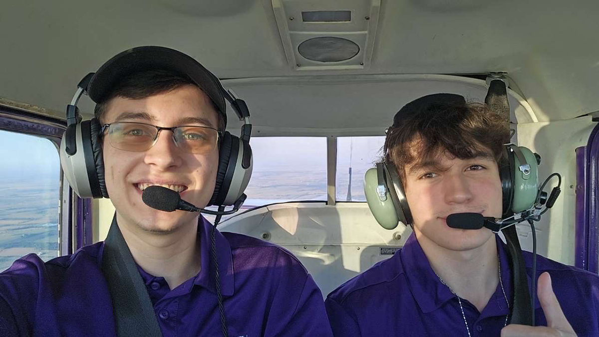 Two male students in purple polos are piloting a plane and taking a selfie-style photo wearing headsets with microphones.