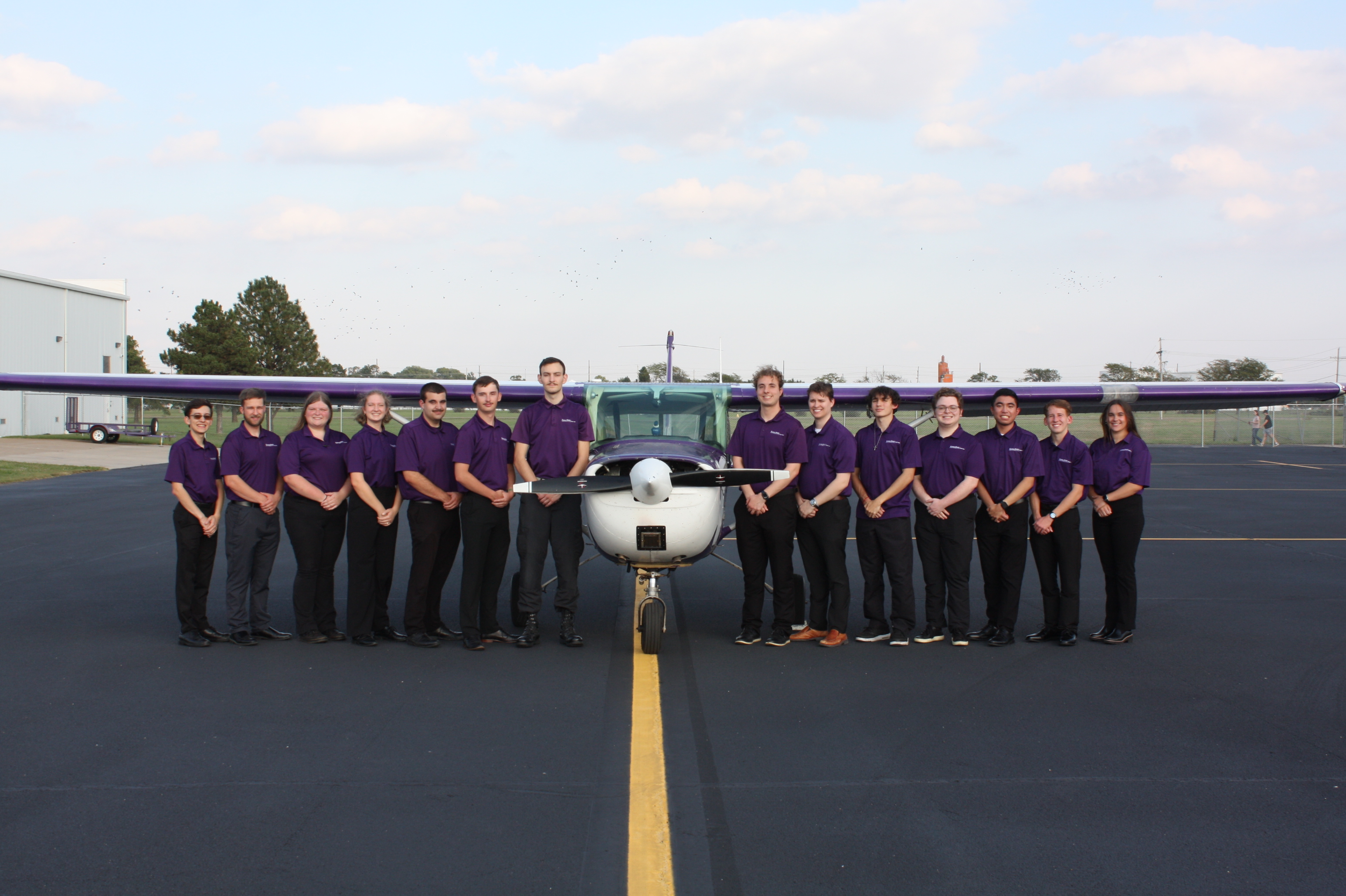 Seven students in purple polos and black pants are pictured in a line on each side of a purple plane with a blue sky in the background.