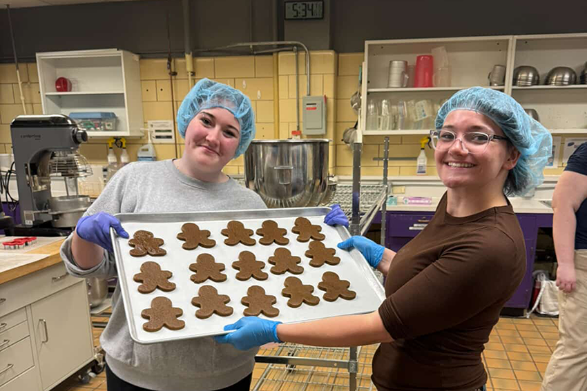 Two students wearing blue hairnets hold up a silver tray of gingerbread people in an industrial kitchen.