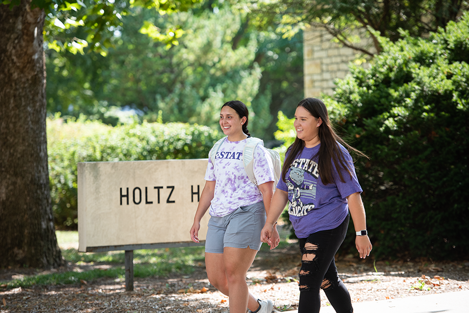 Two female college students walk on campus on a sunny day in front of a limestone sign that says "Holtz Hall."