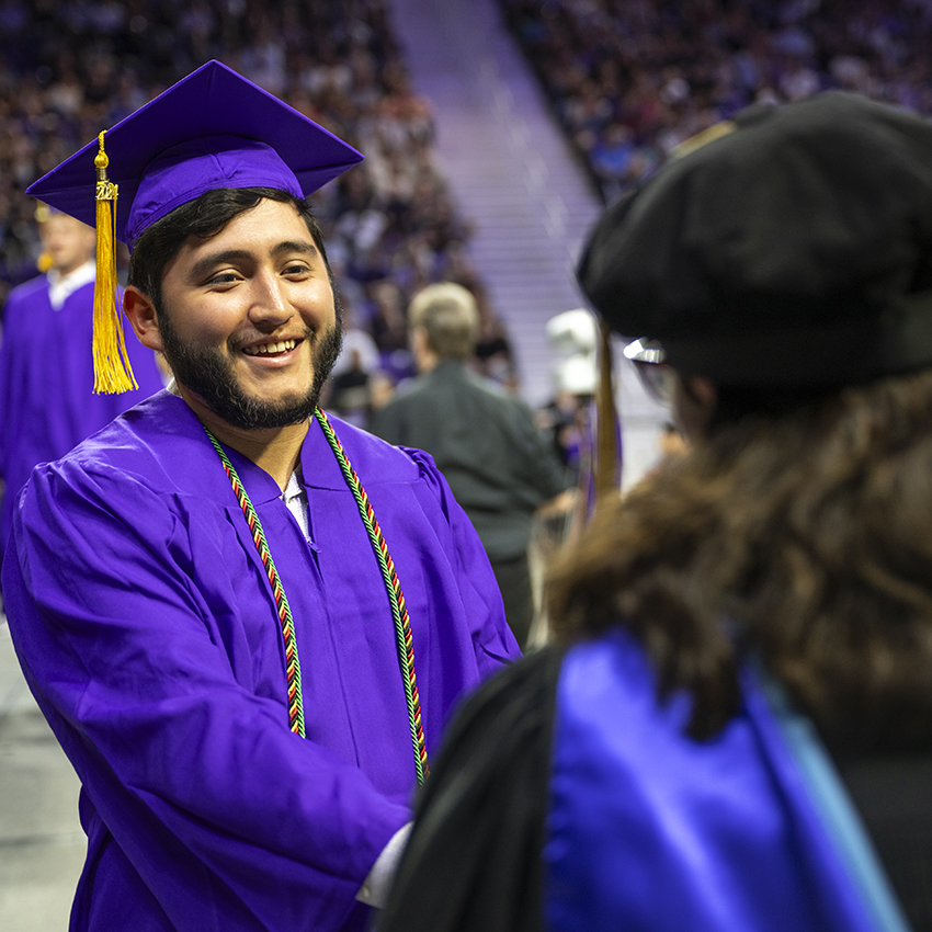 A student in a purple graduation cap and gown shakes the hand of a professor.