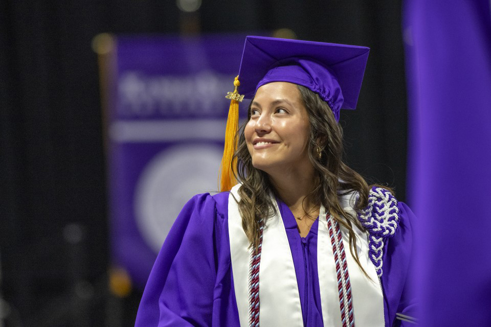 A graduate in a purple cap and gown with a white sash looks over her shoulder and smiles.