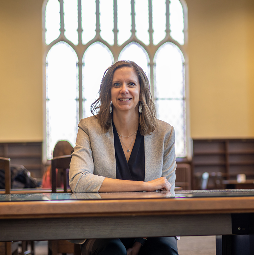 A woman sits at a table in a library and smiles for a portrait.