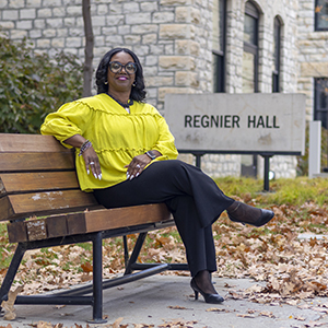 A woman in a yellow blazer sits on a bench outside a limestone college building.