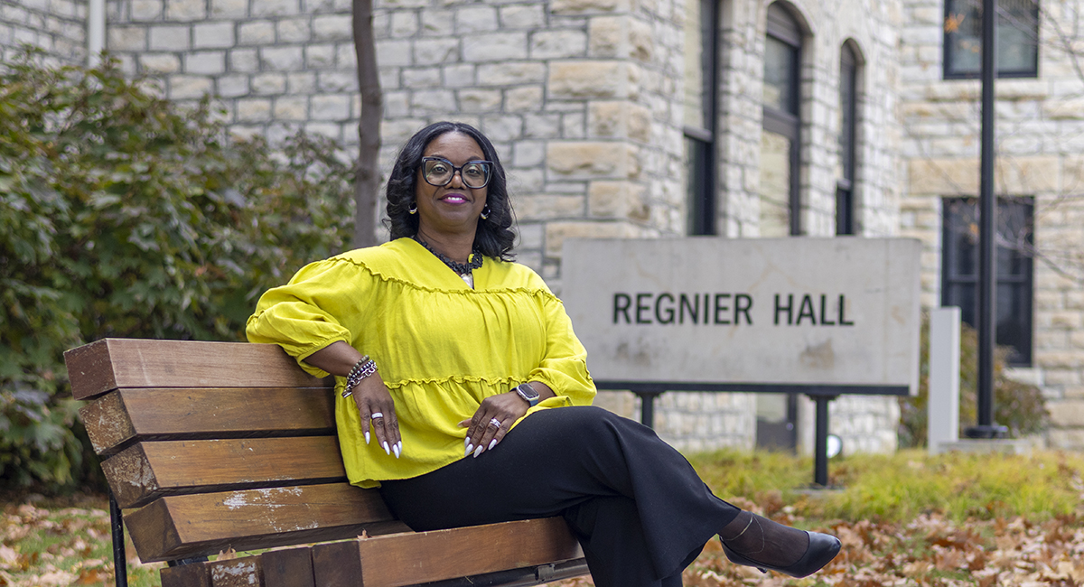 A woman in a yellow blazer sits on a bench outside a limestone college building.