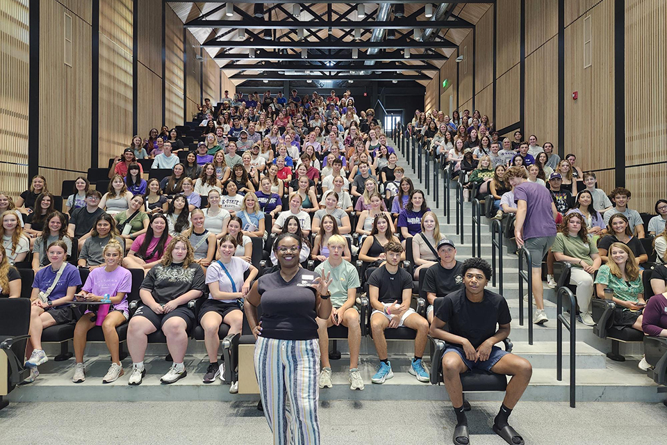 A college student advisor stands at the front of a lecture hall full of college students.