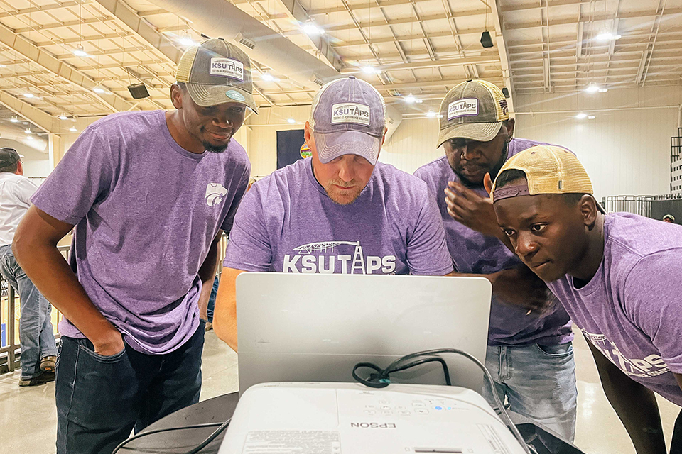 A group of college graduate students crowd around their professor as he uses a computer inside a large barn.