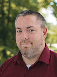 A man with short gray hair is wearing a dark red polo and looking at the camera against an outdoor backdrop.