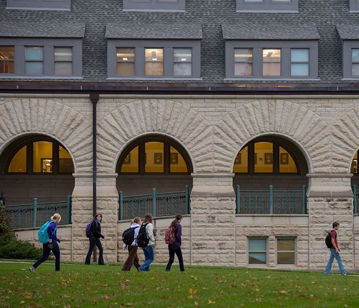 Six college students walk on a cement path through the lawn outside of a limestone library. 