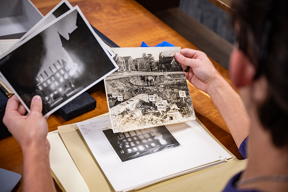 A student wearing sunglasses on the top of their head and a purple "KANSAS STATE UNIVERSITY" shirt sits at a wooden library table and carefuly reads through an old book. A laptop sits next to the book.