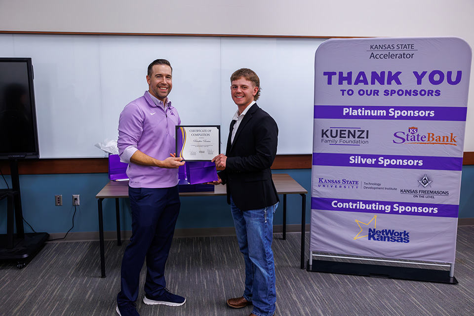 Two people at a K-State Accelerator celebration stand indoors in front of a whiteboard. A man wearing a purple pullover hands a certificate to a young man in a black blazer and jeans.
