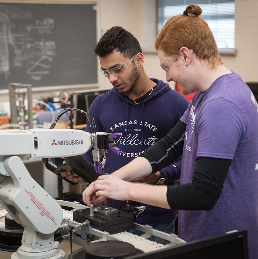 Two Kansas State University students wearing safety glasses work together with a robotic arm and lab equipment during a research activity.