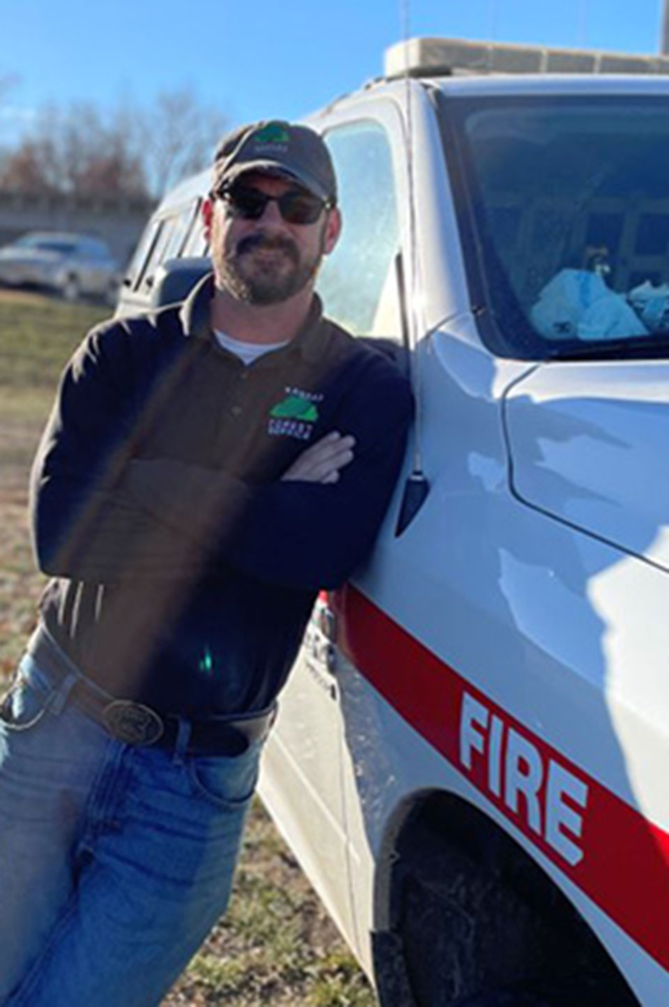 Rodney Redinger wears sunglasses, a black Forest Service polo and stands next to a Forest Service white pick-up truck