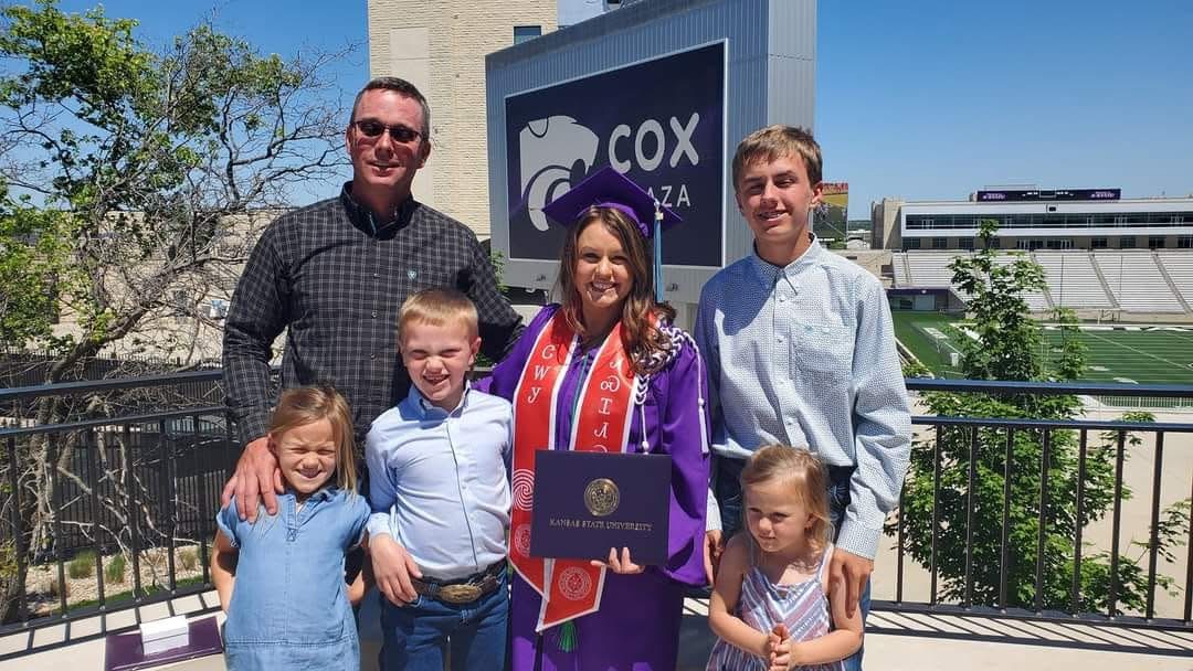 Kelsy Sproul, in a purple cap and gown, holds a Kansas State University diploma folder and stands smiling with her husband and four young children on a balcony at Cox Plaza, with a football stadium in the background on a sunny day.