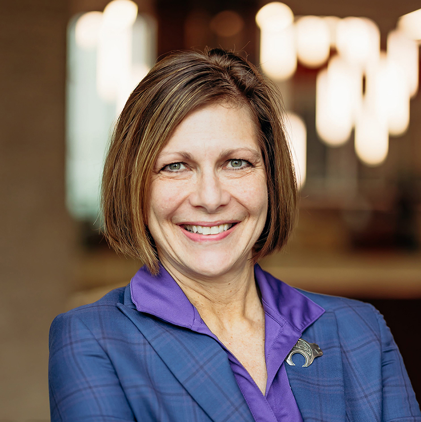 A woman in a blue blazer poses for a portrait in a naturally-lit academic hall.