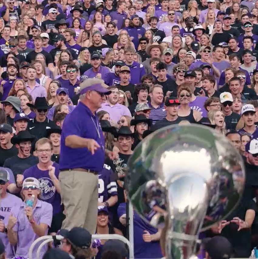 A marching band director stands in front of a student section and a section of tubas at a football game.
