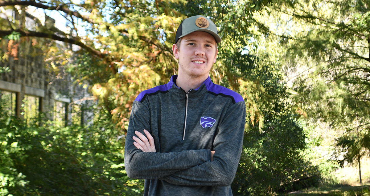 A college student wears a baseball hat and sweatshirt and poses for a portrait outside against a blurry green foliage background. 