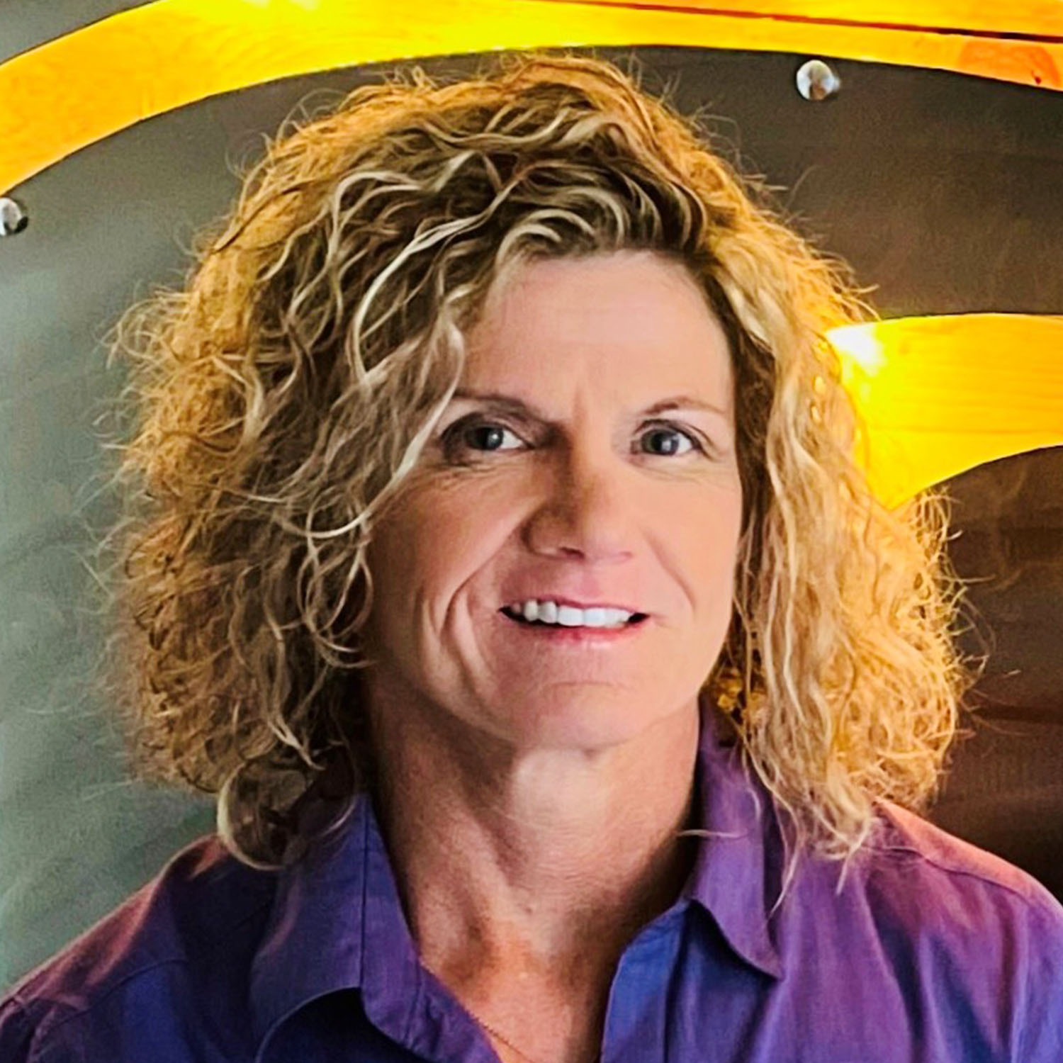 A woman with curly blond hair wearing a purple collared shirt smiles against a dark backdrop with a yellow swirl.