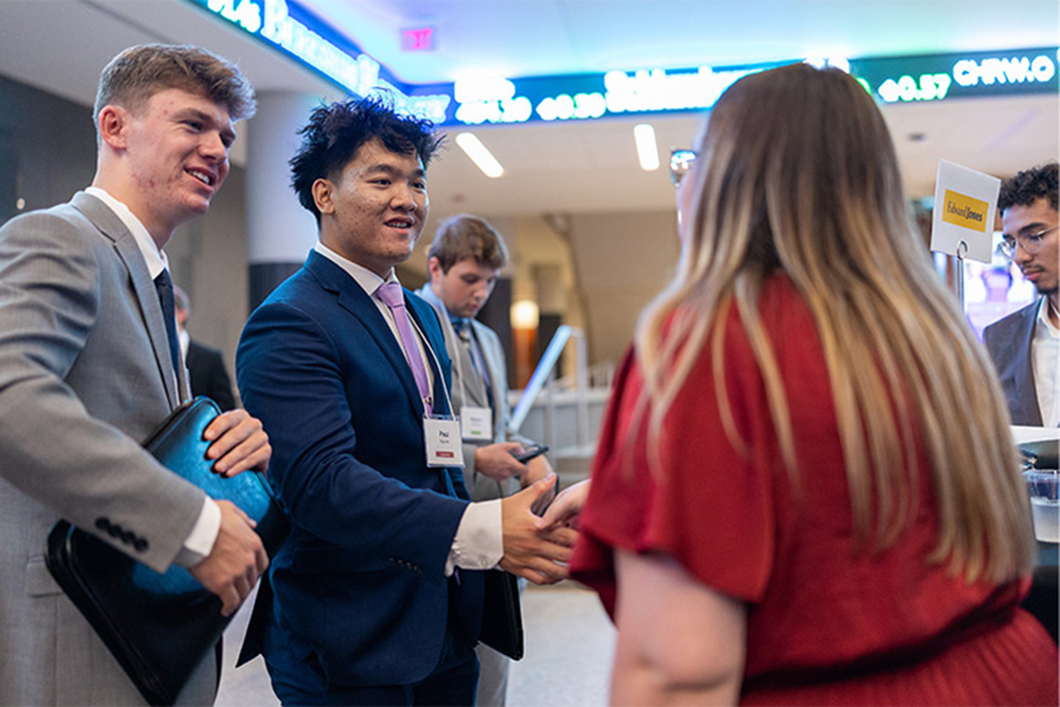 Two students in business attire greet and shake hands with an attendee at a professional networking event inside a campus building.