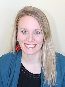 A woman with blonde hair weraing a blue cardigan with orange earrings smiles.