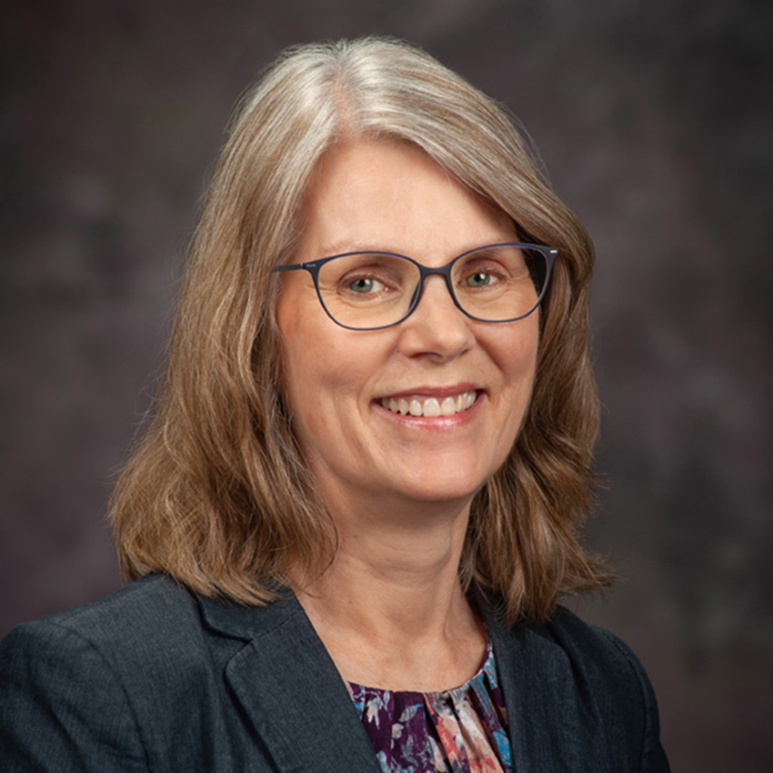 A woman with shoulder-length light brown hair, glasses, and a dark blazer smiles for a professional portrait. 