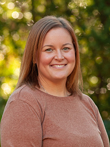 A woman with light brown hair smiles for a photo against a green outdoor background.