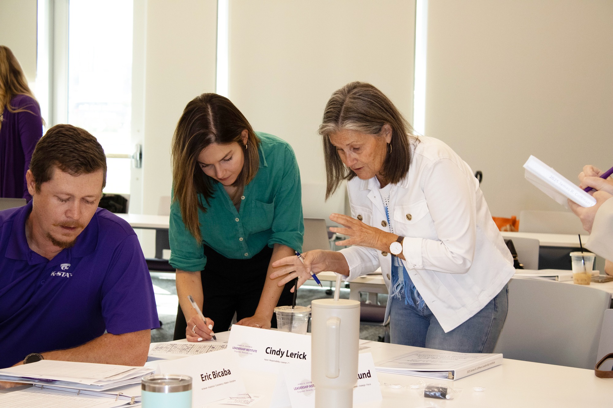 Three adults collaborate at a table during a workshop. A woman in a green shirt leans over to write on a document while a man in a purple K-State polo reviews papers beside her. Another woman in a white jacket gestures as she offers guidance. Name tents and notebooks are visible on the table.