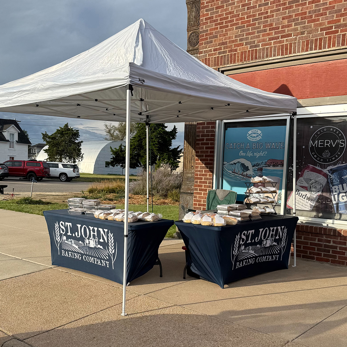 Two tables of baked goods sit outside in front of a brick building and under a white popup tent.