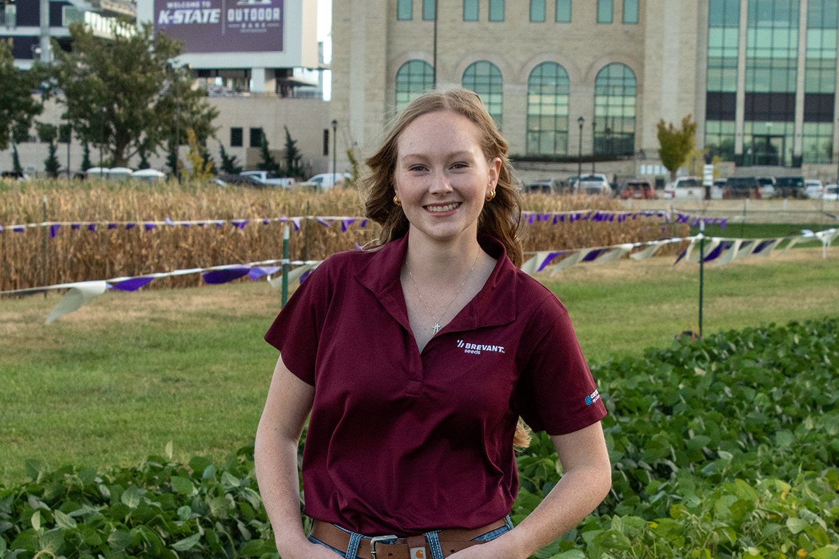 A woman wears a maroon polo shirt and stands outside in a field with a football stadium in the background. 