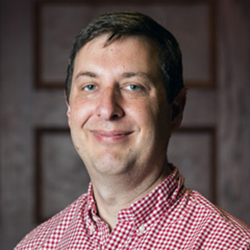 A man with short brown hair and a red-check button-up shirt smiles for a professional portrait. 