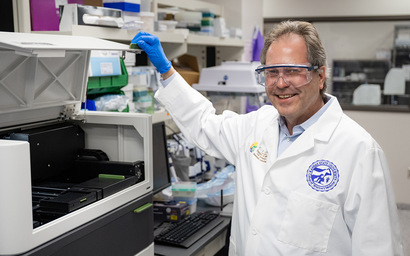 A veterinary researcher wearing safety glasses and a white lab coat poses for a portrait next to an analysis machine in a lab.