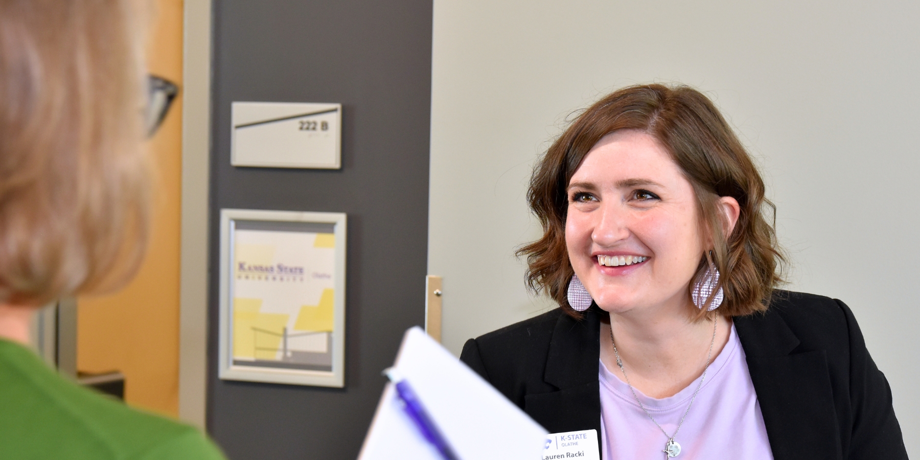 Lauren Racki smiles while speaking to a colleague. She is wearing a purple blouse and a black blazer.
