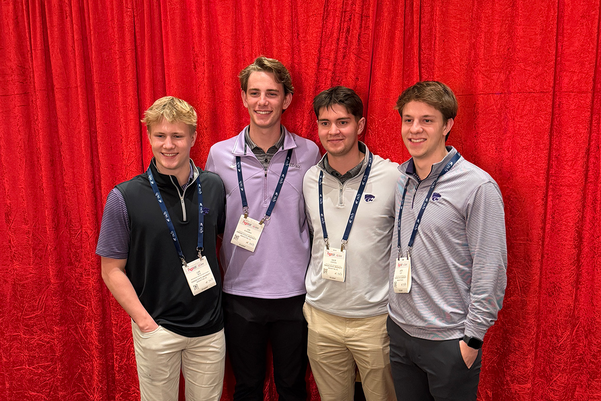 Four college students wearing business casual attire and event lanyards stand in front of a bright red cloth backdrop for a group photo. 