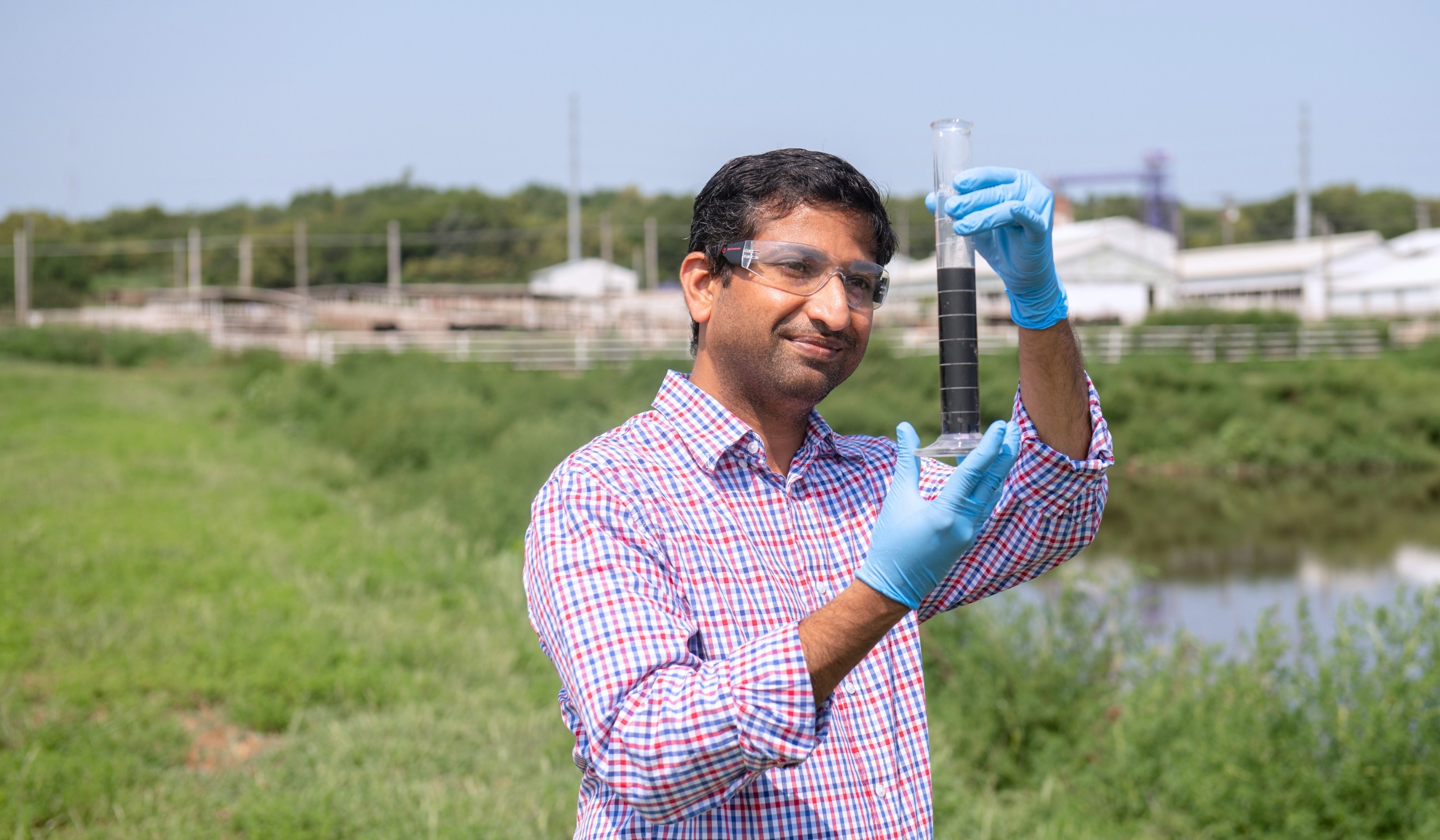 Prathap Parameswaran, wearing lab goggles, a long-sleeve button-up shirt and blue latex gloves, holds up a rain gauge to inspect its contents. In the blurred background, there is a grassy area, a small body of water and a farming operation.