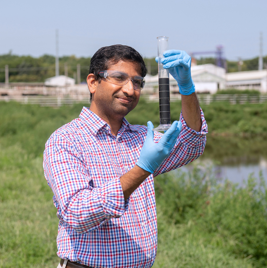 Prathap Parameswaran, wearing lab goggles, a long-sleeve button-up shirt and blue latex gloves, holds up a rain gauge to inspect its contents. In the blurred background, there is a grassy area, a small body of water and a farming operation.