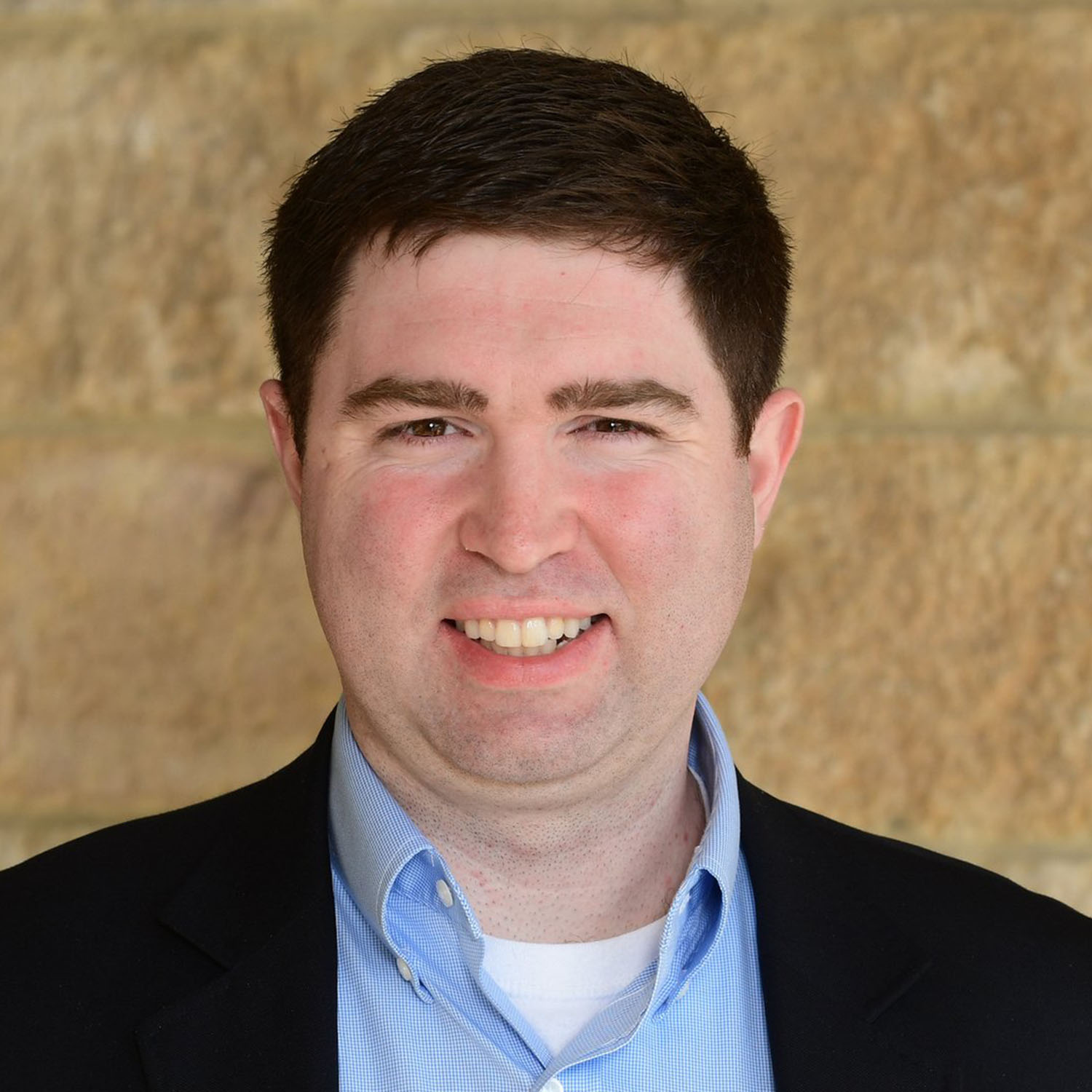 A man with dark brown hair wearing a black blazer and light blue collared shirt smiles for a portrait photo.