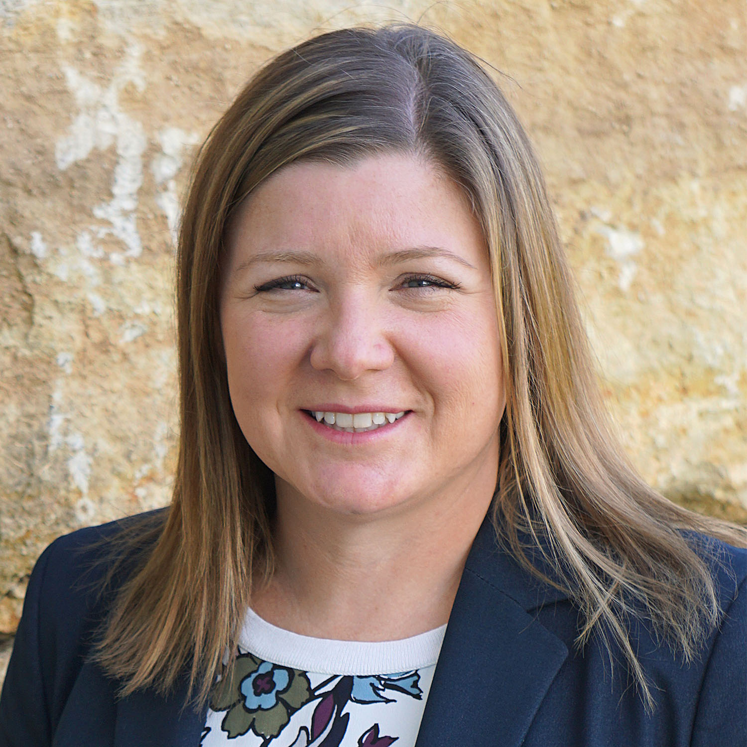 A woman with light brown, shoulder-length hair wearing a navy blazer and a white shirt with flowers on it smiles against a stone background.