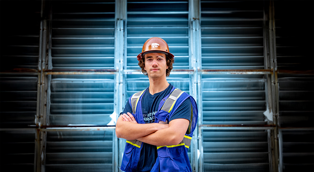 A male student wearing a black t-shirt and purple safety vest stands against a garage door illuminated in a blue glow.