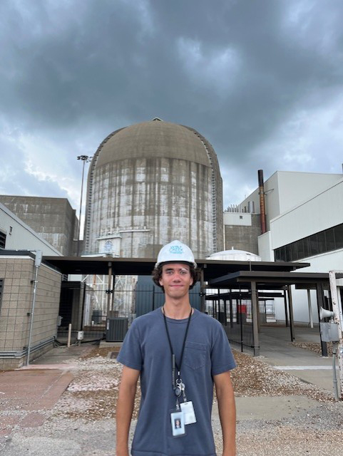 A male intern in a blue t-shirt and a white hard hat stands in front of the cement dome of Wolf Creek Nuclear Generating Station.