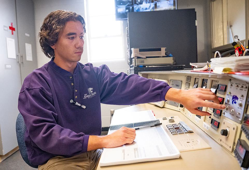A young man with brown hair wearing a long-sleeved purple shirt points at an instrumentation panel on the desk in front of him behind a notebook.