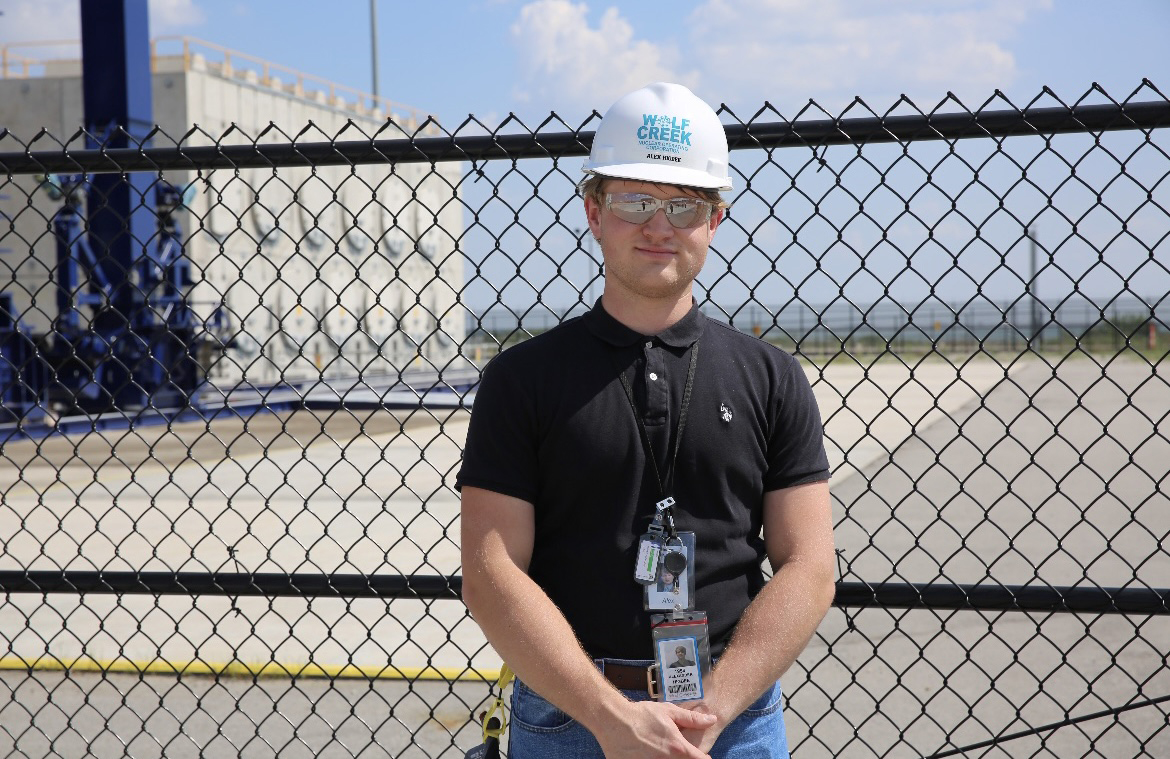 A white man in a black polo wears a white hard hat that says Wolf Creek on it in blue letters. He stands in front of a black fence, with a concrete structure visible in the background.