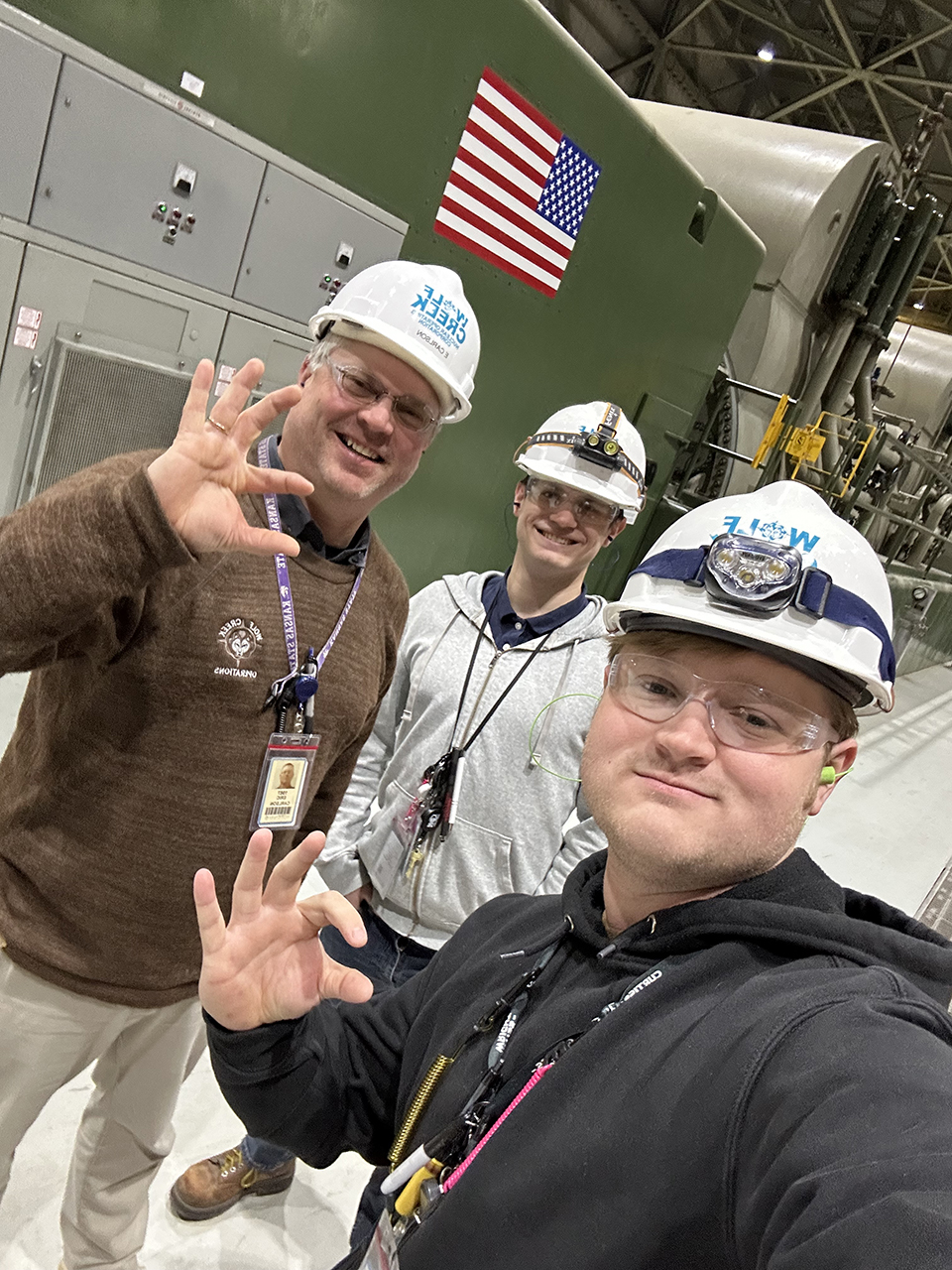 Three men in white hardhats are pictured with a large green piece of equipment with an American flag on it. Two of the men are holding up Wildcat hand signs.
