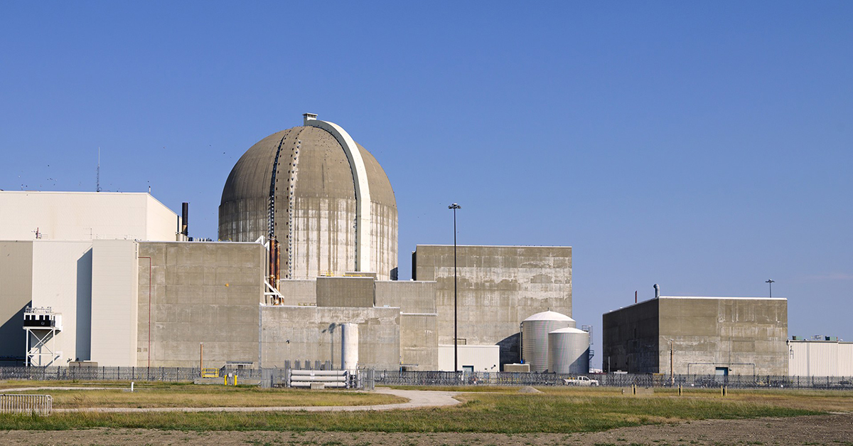 Wolf Creek Nuclear Generating Station is pictured against a clear blue sky. The parts of Wolf Creek shown include square and rectangular cement buildings as well as a large cement dome. Some grass is seen in the foreground.