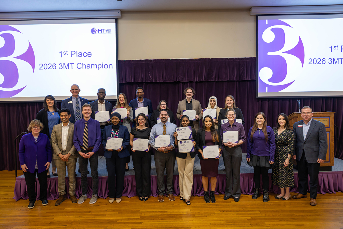 A large group of graduate students on a platform hold certificates alongside a number of expert judges at a research presentation competition in a ballroom.